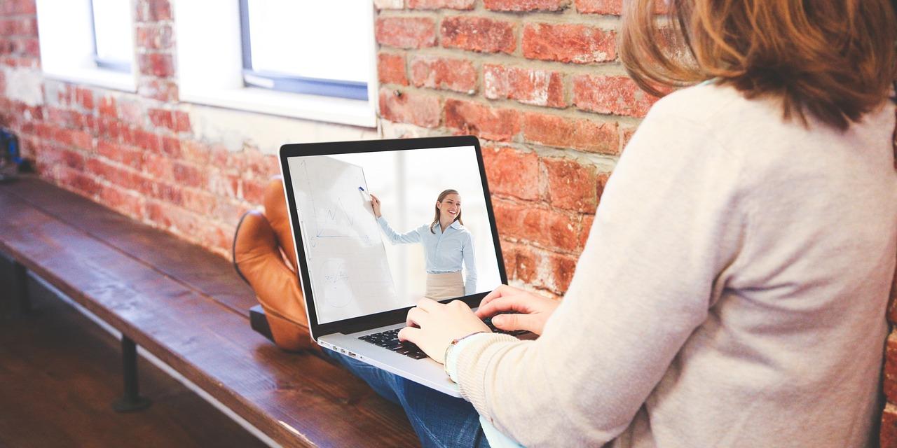 Eine Person sitzt mit Laptop auf einer Bank, auf dem Bildschirm sieht man eine referierende Person