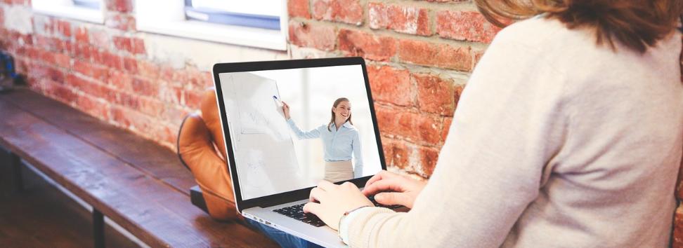 Eine Person sitzt mit Laptop auf einer Bank, auf dem Bildschirm sieht man eine referierende Person