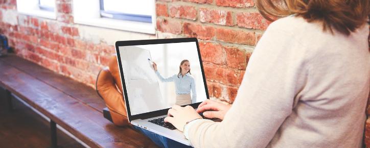 Eine Person sitzt mit Laptop auf einer Bank, auf dem Bildschirm sieht man eine referierende Person